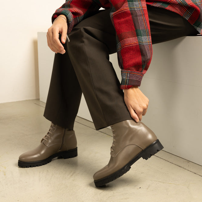 Person wearing olive green leather boots and a red plaid shirt sitting on a white wall.