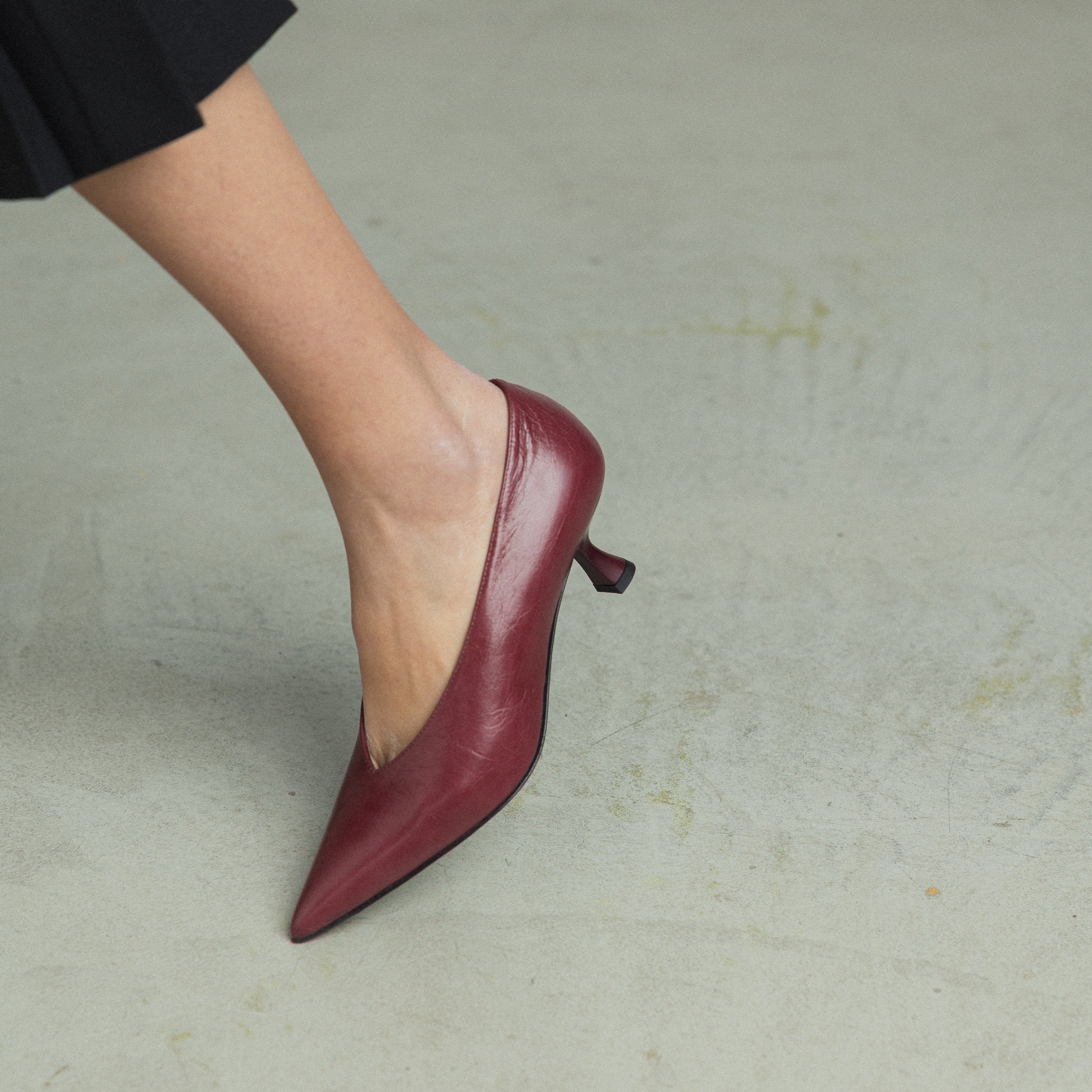 A close-up of a person's foot wearing a pointed-toe, mid-heel mule shoe in a deep burgundy red, paired with a black skirt, standing on a textured light gray concrete floor.