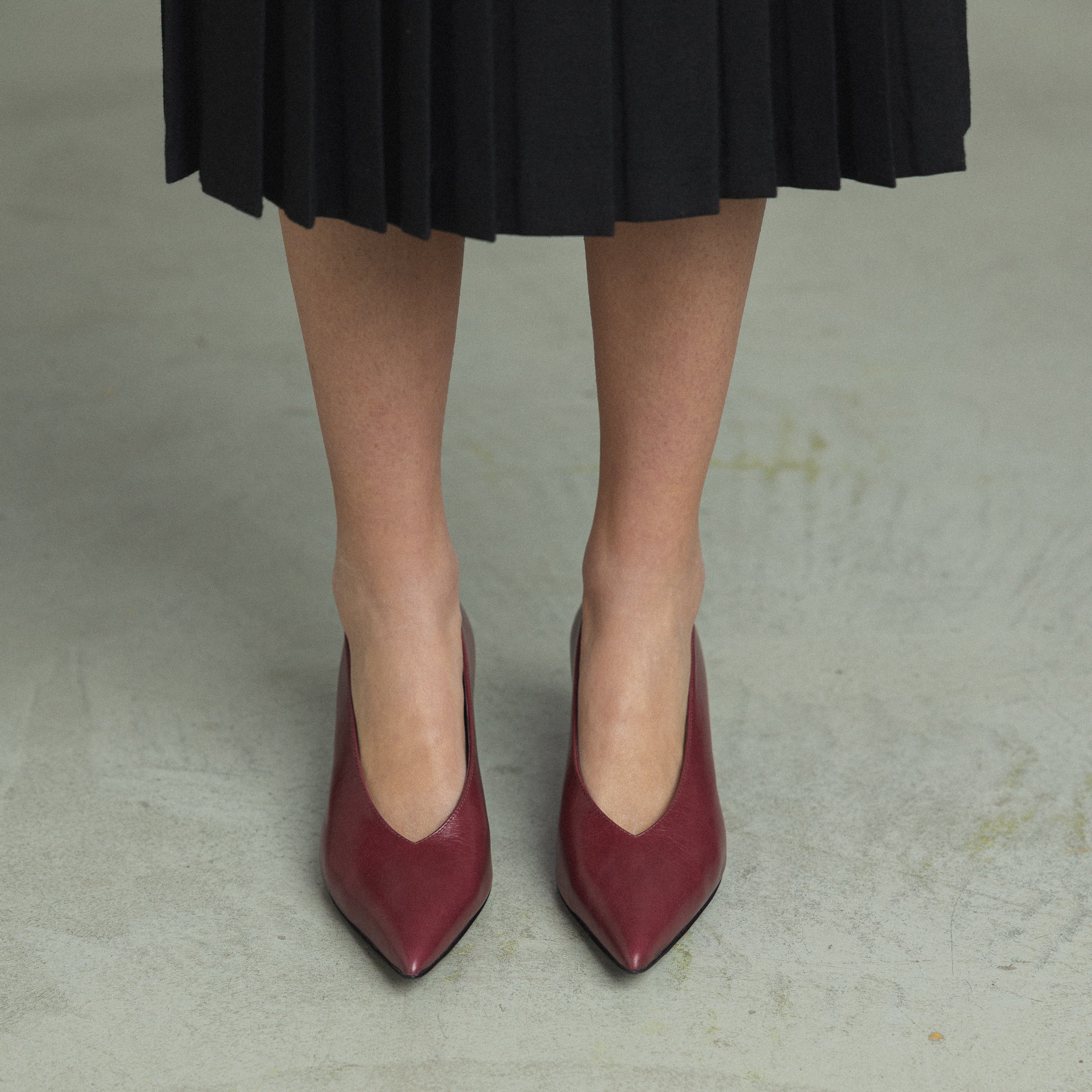 A person wearing a black pleated skirt and burgundy pointed-toe pumps, standing on a light gray concrete floor.