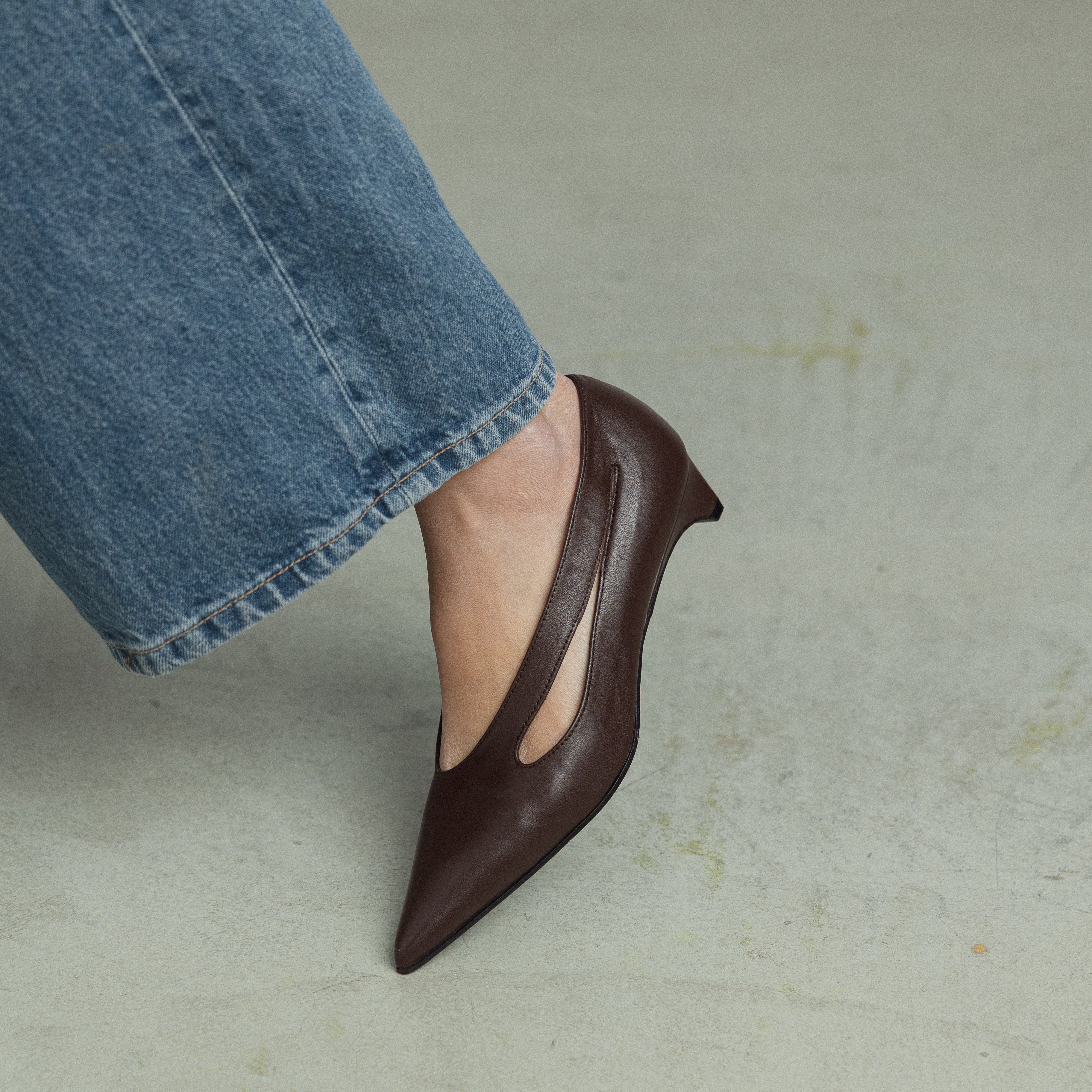 A person wearing blue jeans and a dark brown pointed-toe flat shoe with a side cutout, standing on a light gray concrete floor.