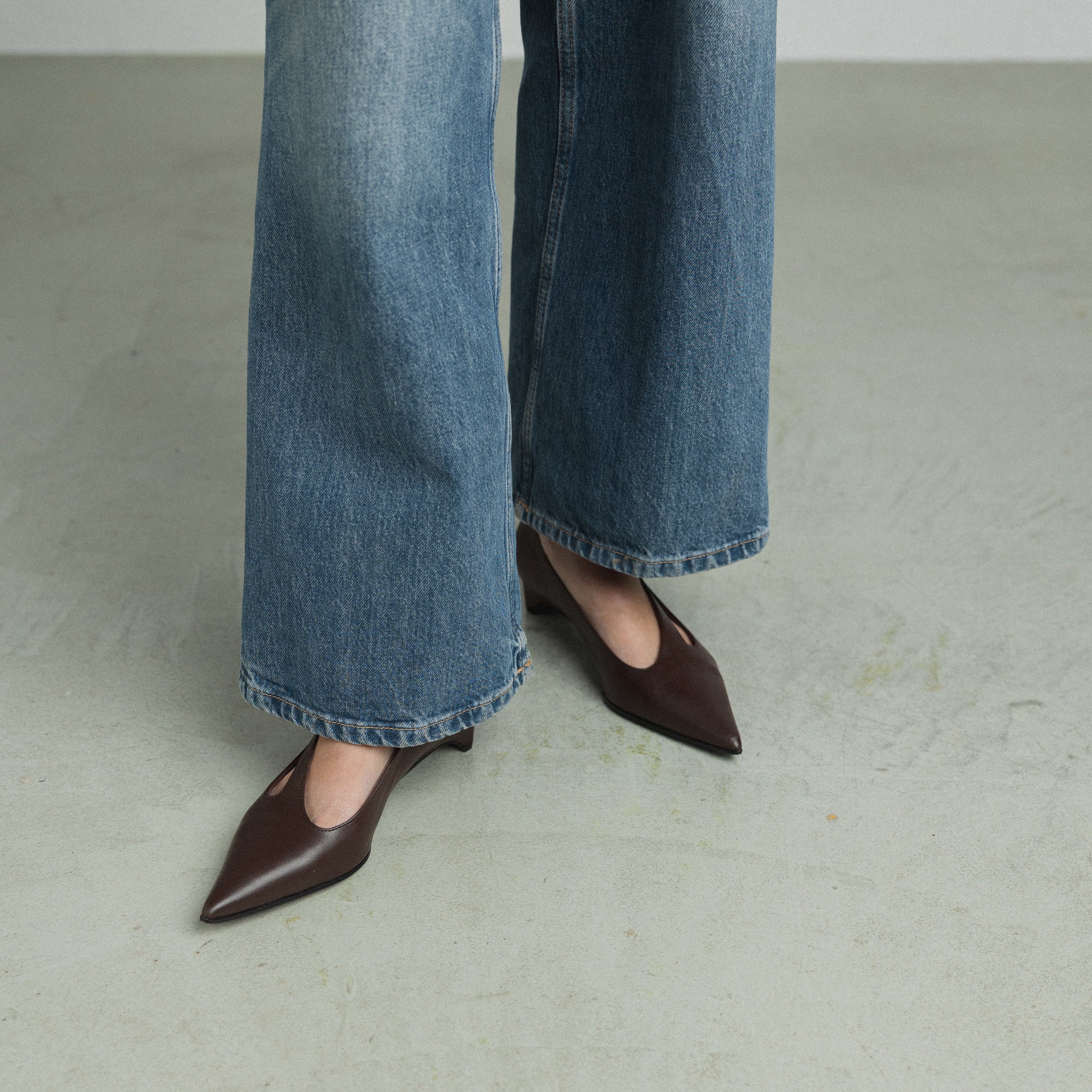 A person wearing blue wide-leg jeans and dark brown pointed-toe flats, standing on a light gray concrete floor.