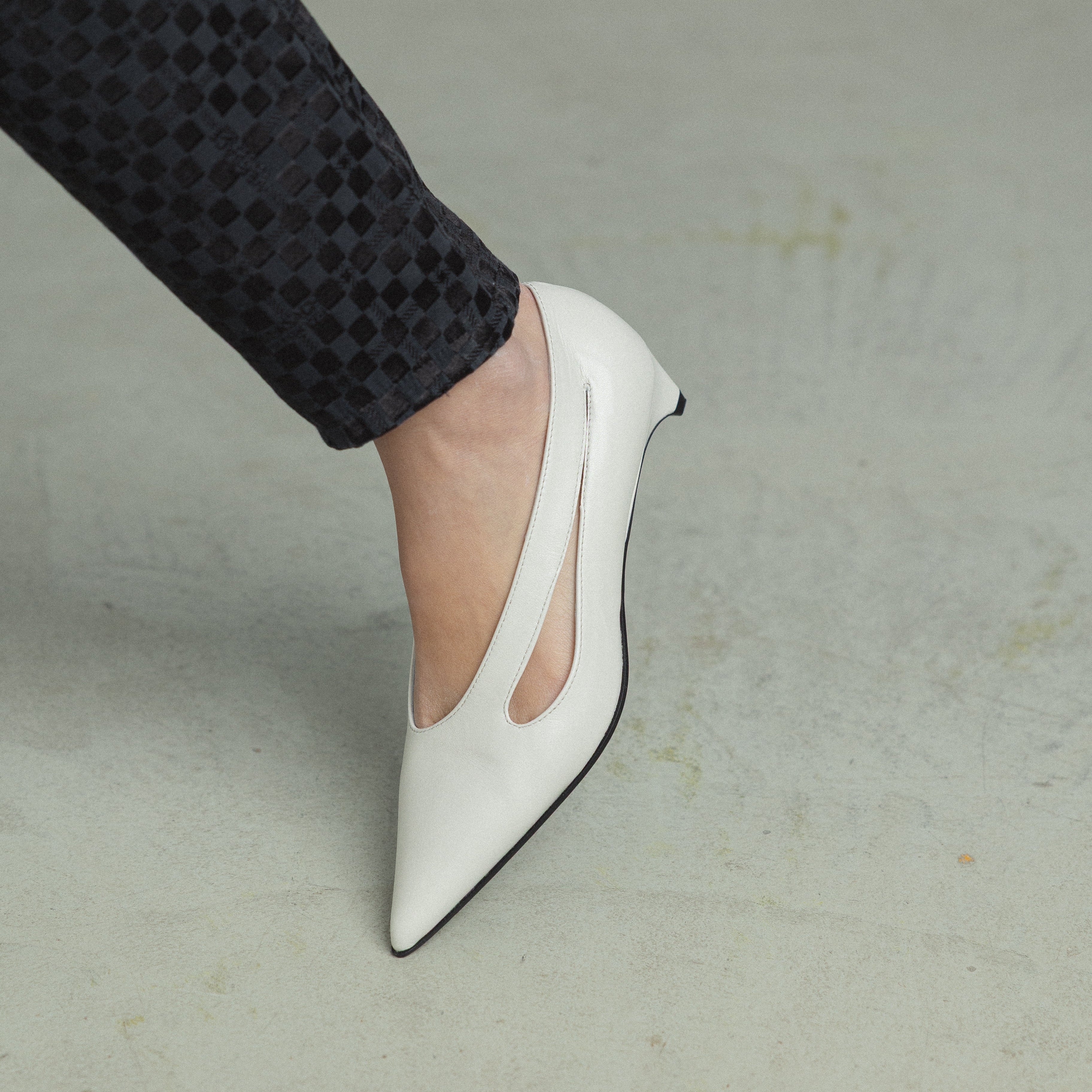 A close-up of a person's foot wearing a white pointed-toe pump with a side cutout, paired with black patterned trousers, standing on a light gray concrete floor.