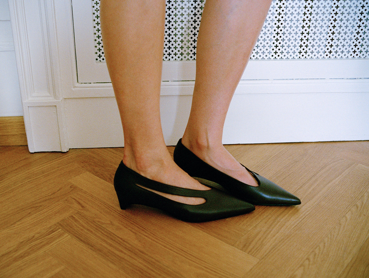 A person wearing black pointed-toe kitten-heel pumps with a cutout detail, standing on a light wood herringbone floor in front of a white baseboard and patterned wall.